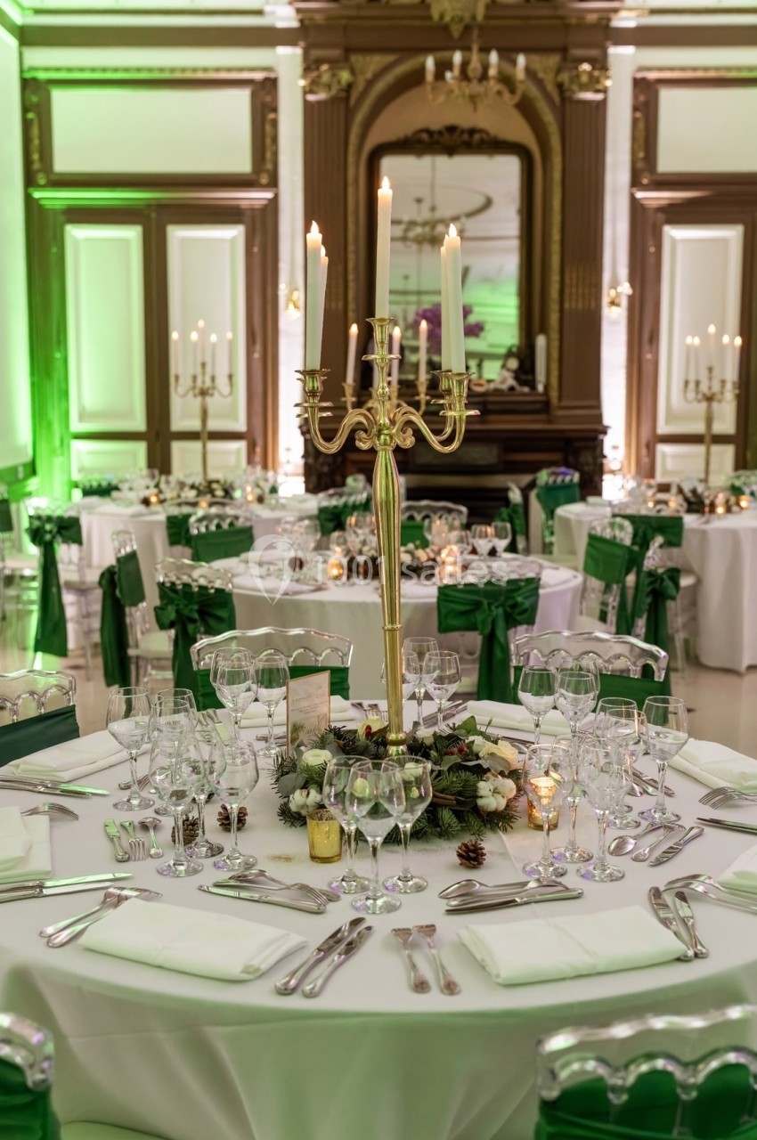 Salle de réception élégante avec tables décorées, nappes blanches, chandeliers dorés et éclairage vert.