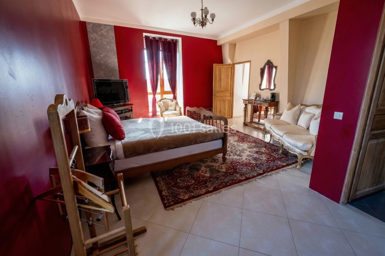 Chambre spacieuse avec murs rouges, lit double, canapé, tapis oriental, et lumière naturelle entrant par une fenêtre.
