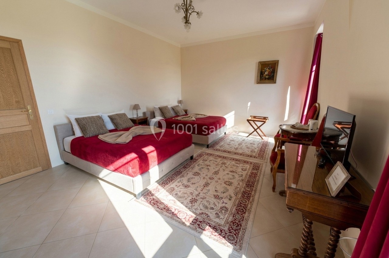 Chambre lumineuse avec deux lits doubles, tapis au sol, bureau en bois et rideaux rouges encadrant une fenêtre.