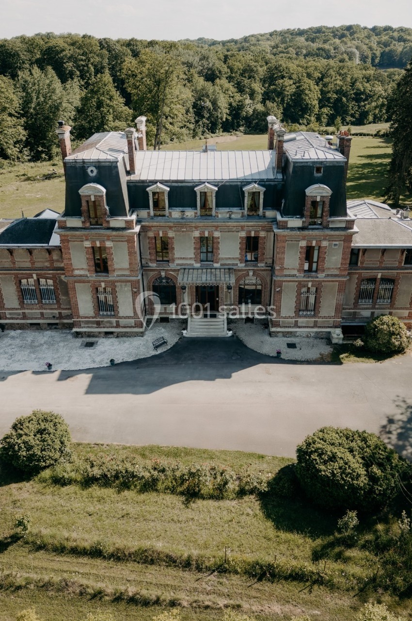 Vue aérienne d'un château en briques rouges avec toit mansardé, entouré de verdure et d'une allée.