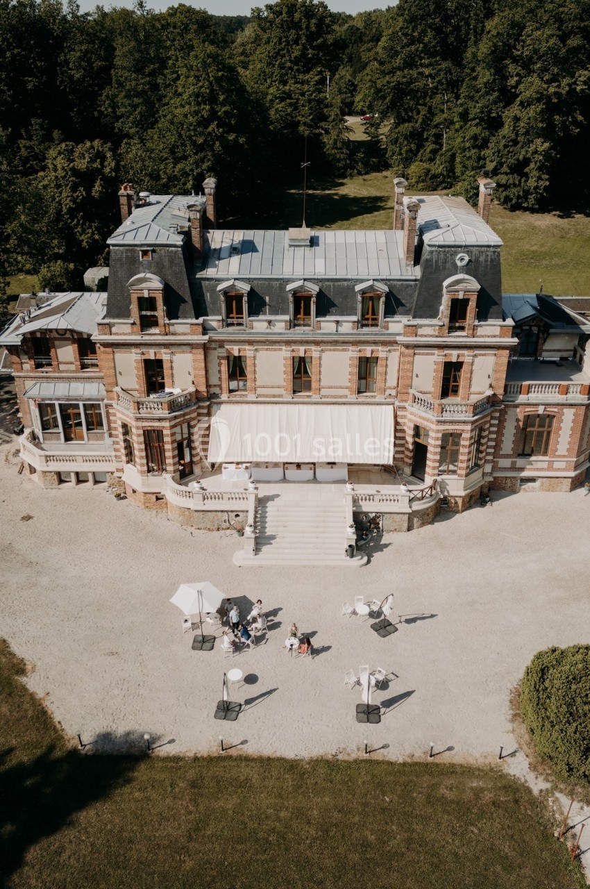 Vue aérienne d'un château en briques entouré de verdure, avec une terrasse aménagée et des parasols au premier plan.