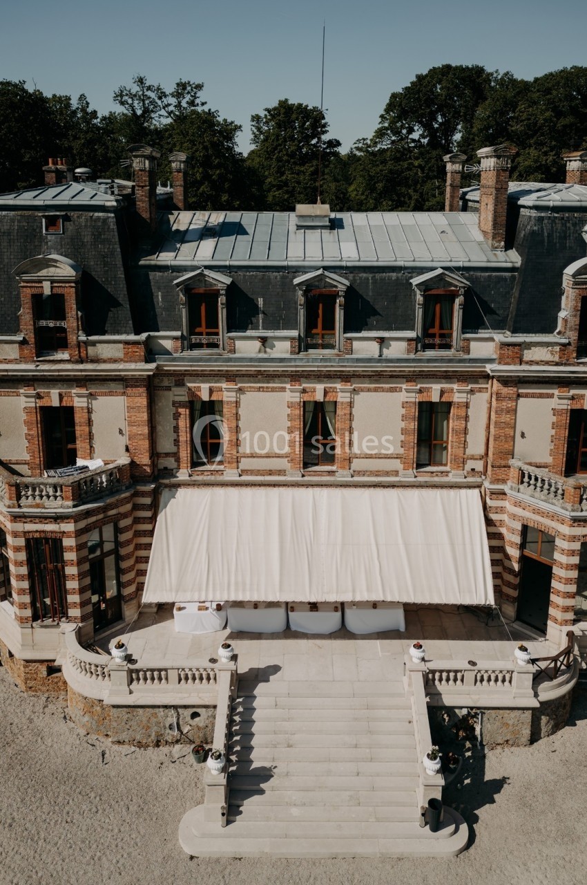 Façade d'un château en briques et pierre avec escalier central, terrasse et auvent beige, entouré de végétation.