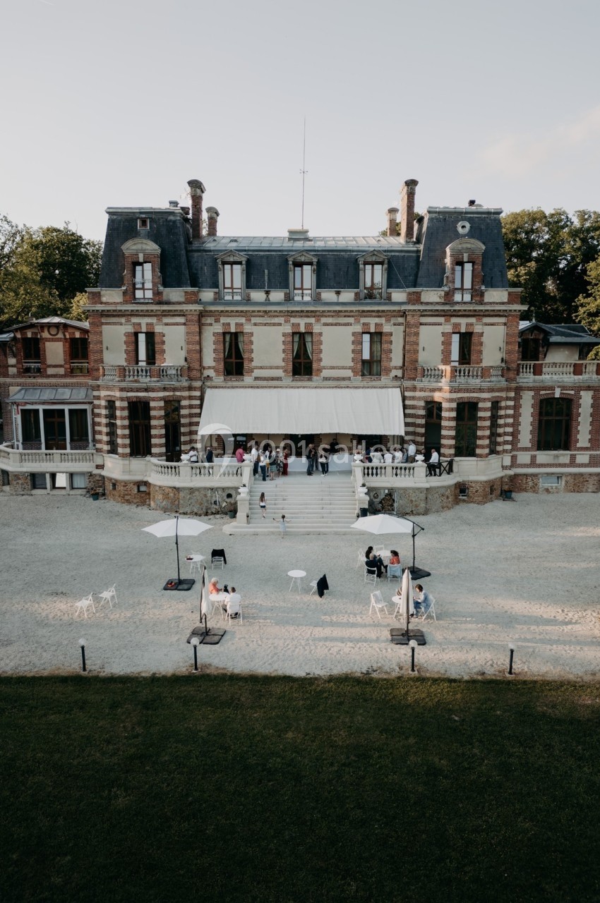 Façade d'un grand bâtiment en briques avec toit mansardé, terrasse aménagée et personnes rassemblées devant.