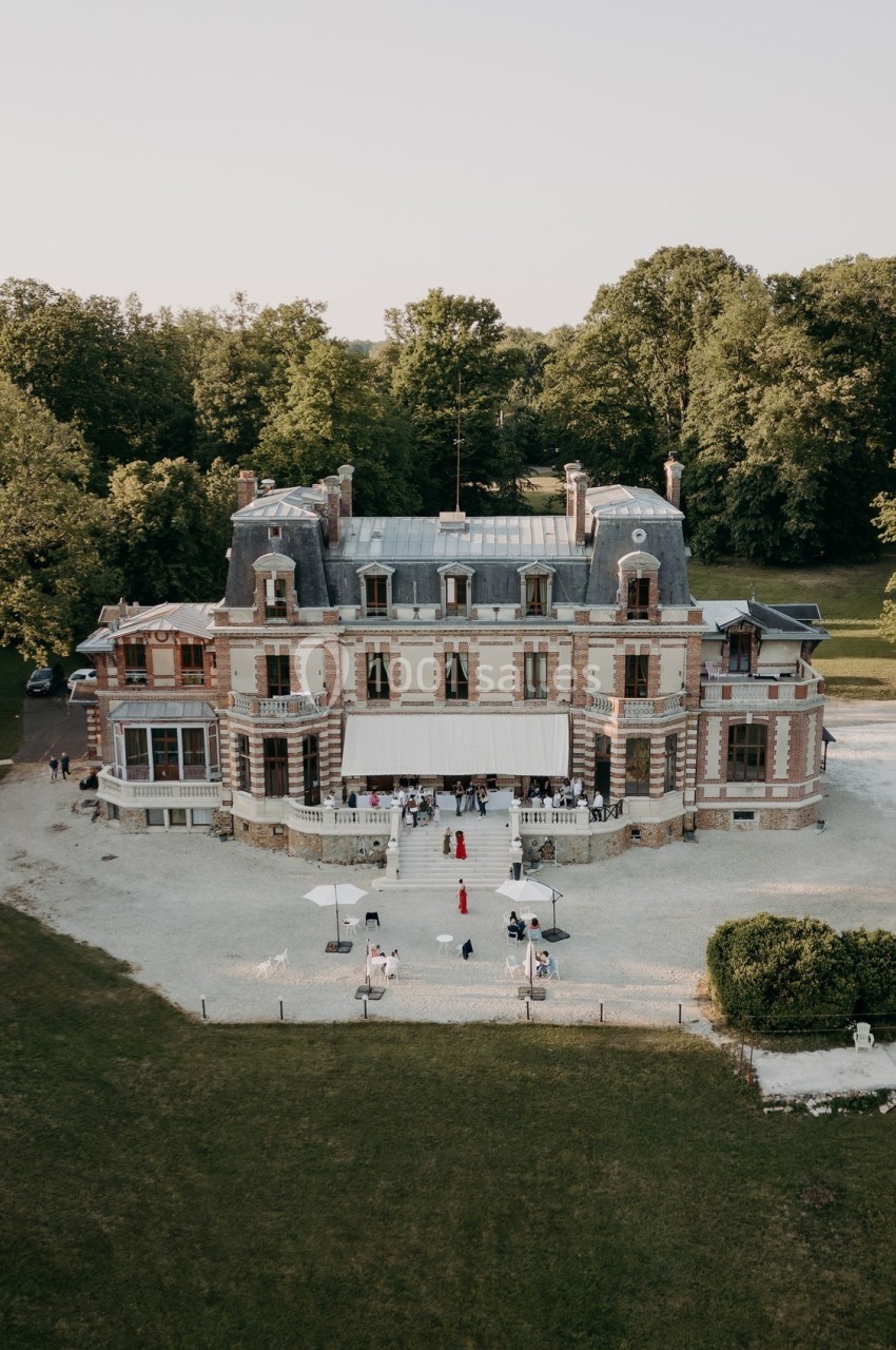 Vue aérienne d'un château en briques entouré de verdure, avec des invités rassemblés sur le parvis.