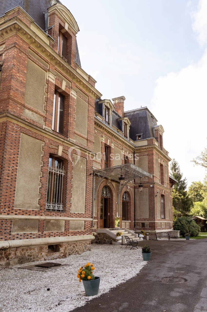 Façade d'un bâtiment en briques rouges avec toit mansardé, entouré de végétation et décoré de pots de fleurs.