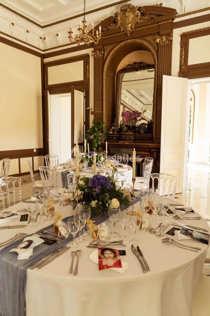 Salle élégante avec table ronde dressée, décorée de fleurs et bougies, entourée de chaises transparentes.