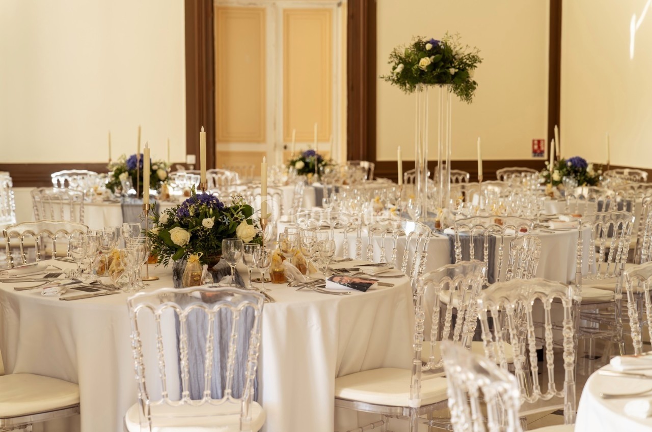 Salle de réception décorée avec des tables rondes, nappes blanches, chaises transparentes et centres de table fleuris.