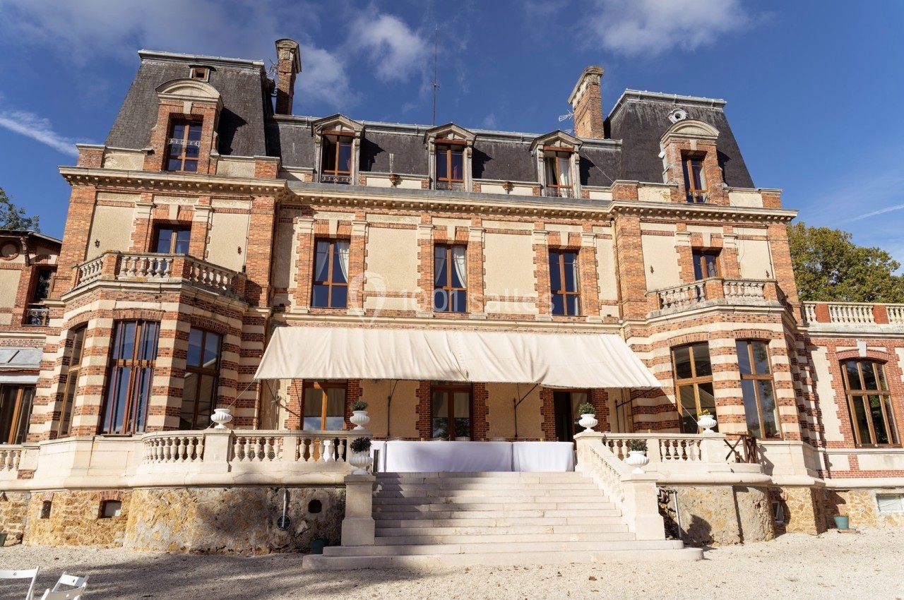 Façade d'un château en briques et pierres avec balcons, grandes fenêtres et une terrasse surmontée d'une toile beige.