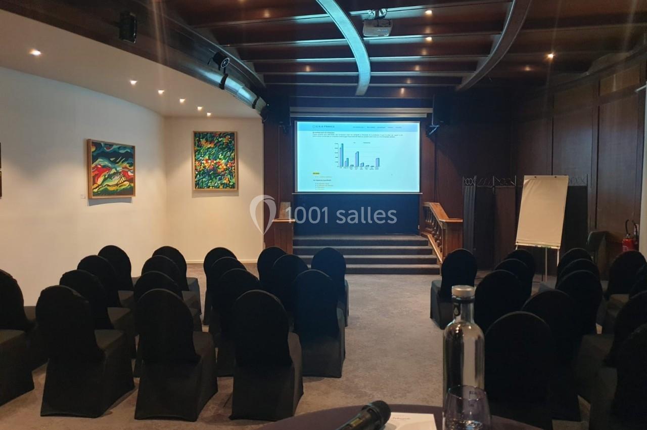 Salle de conférence vide avec des chaises noires, un écran de projection allumé, des tableaux accrochés et un chevalet blanc.