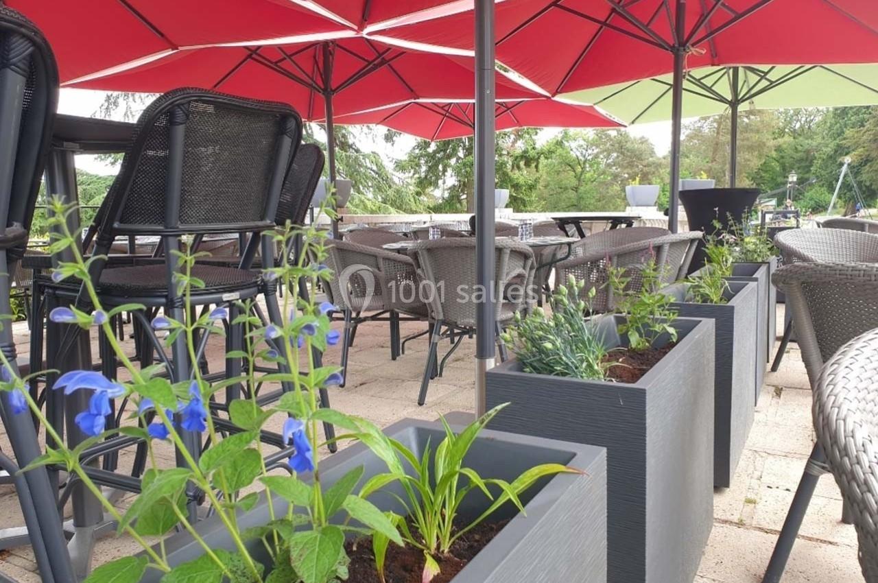 Terrasse aménagée avec tables, chaises en osier, parasols rouges et jardinières contenant des plantes fleuries.