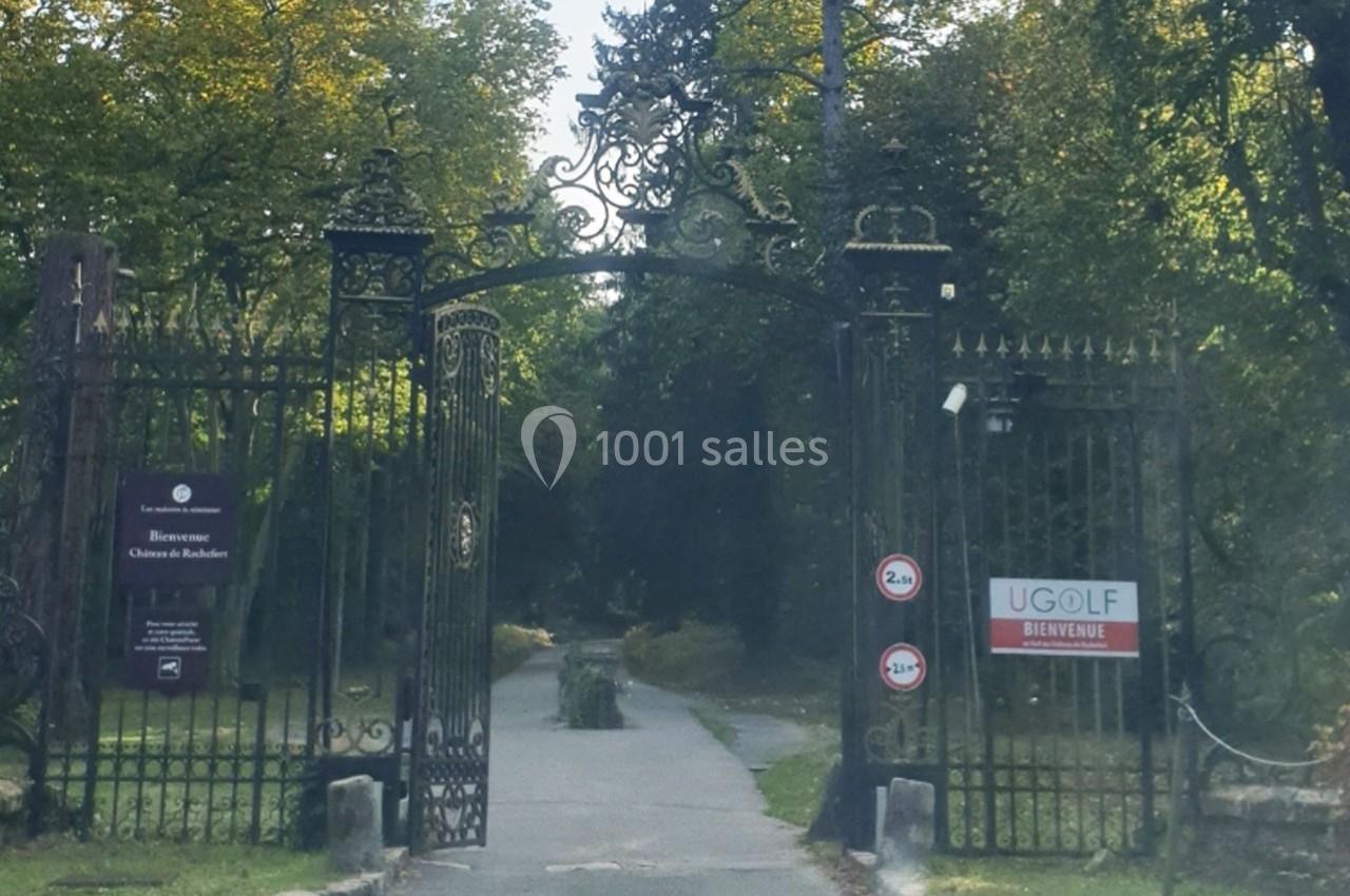 Portail en fer forgé ouvrant sur une allée bordée d'arbres, avec des panneaux d'accueil et de signalisation.