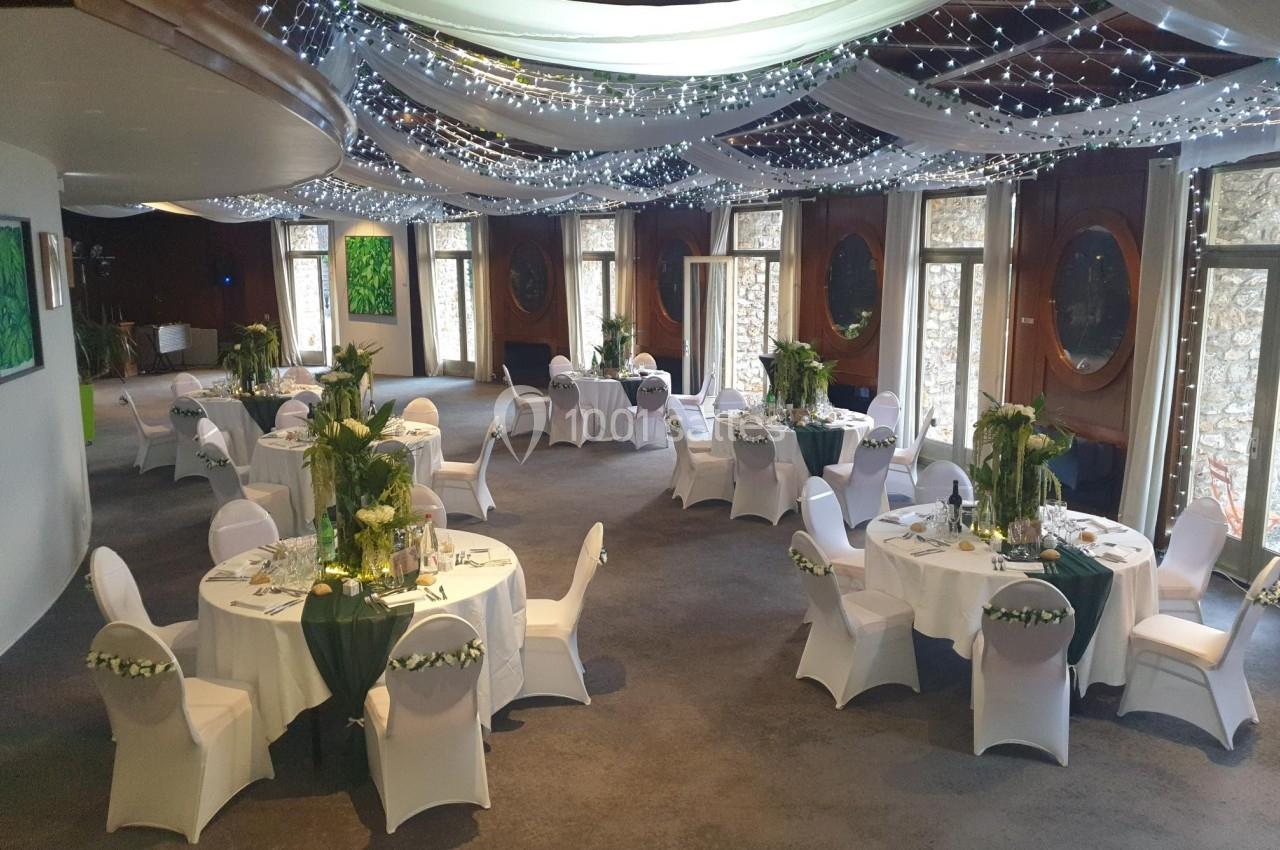 Salle de réception décorée avec des tables rondes, nappes blanches, centres de table verts et guirlandes lumineuses…