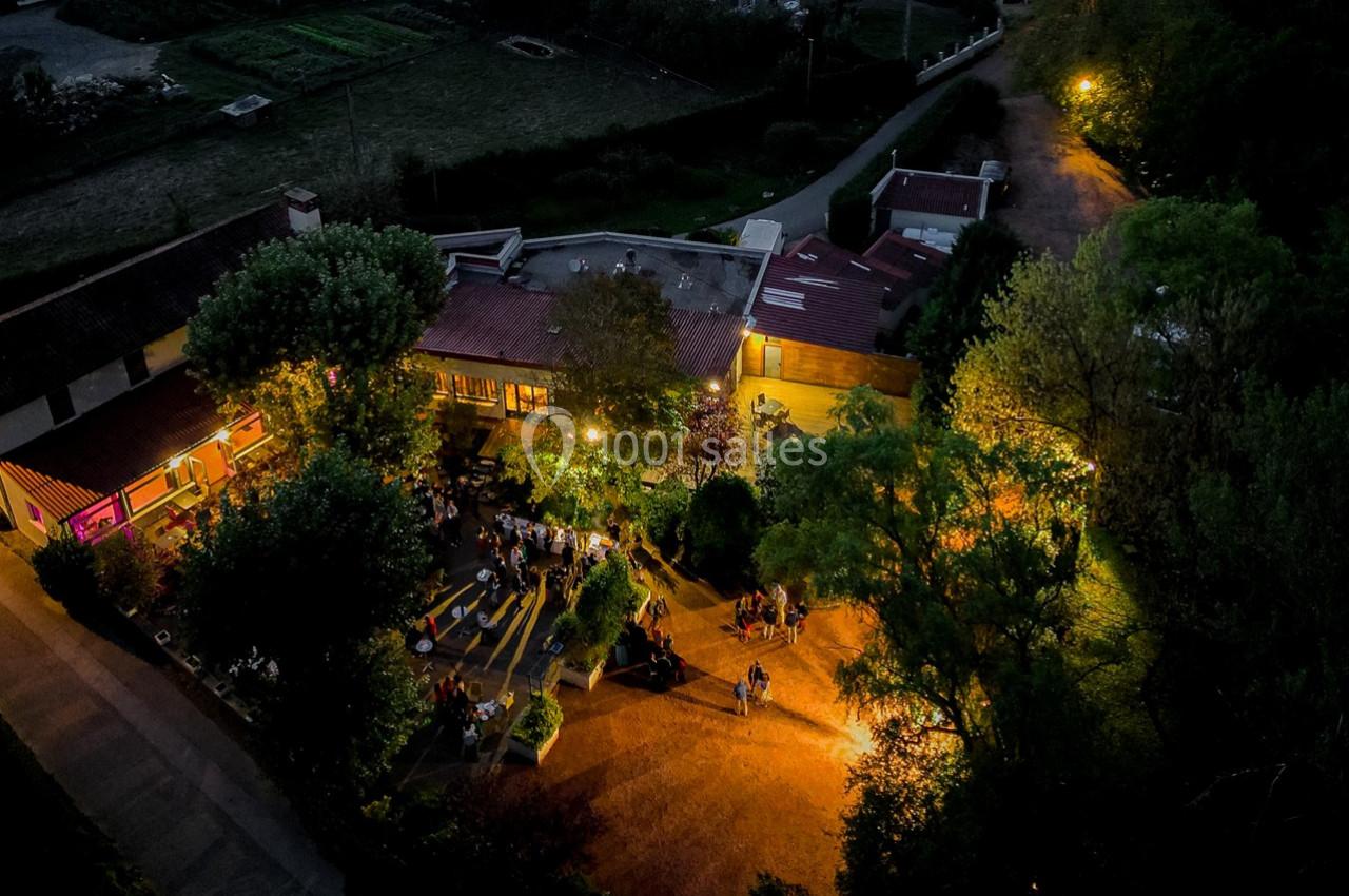 Vue aérienne nocturne d'un espace extérieur éclairé avec des personnes rassemblées près d'un bâtiment entouré de végétation.