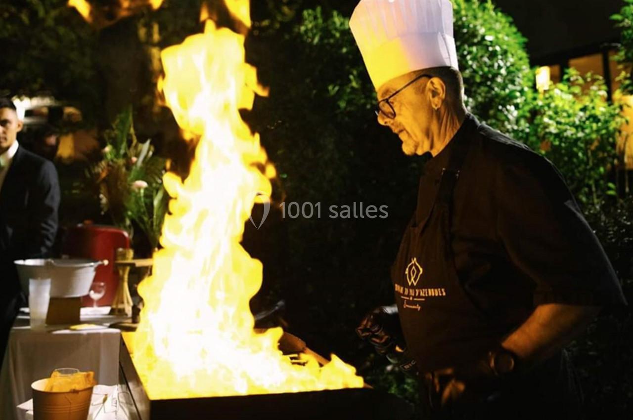 Un chef en toque blanche cuisine sur une plaque chauffante avec des flammes vives dans un cadre extérieur nocturne.