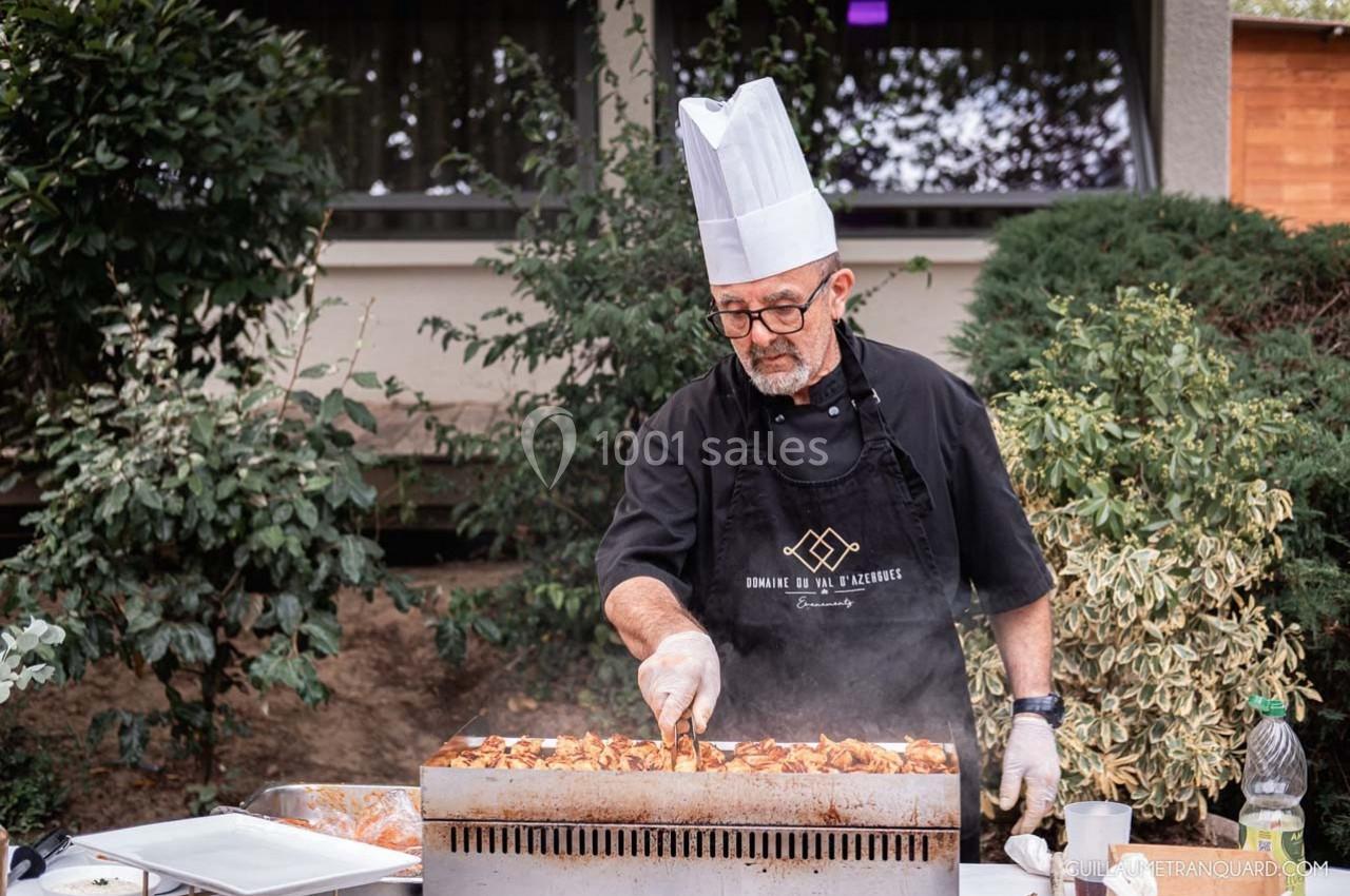 Un chef en tablier et toque cuisine des légumes sur une grande plaque chauffante en extérieur.