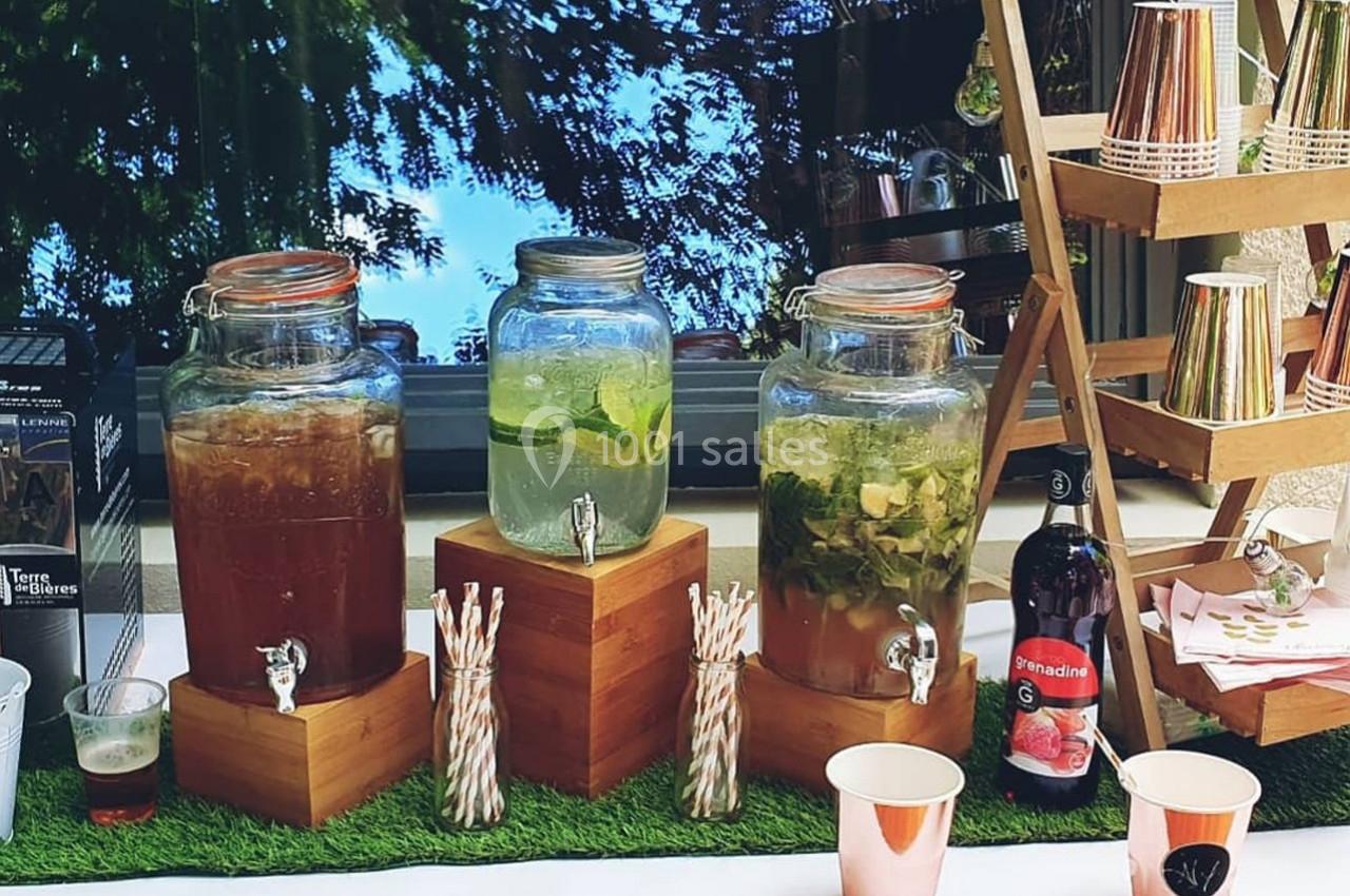 Trois distributeurs de boissons avec pailles, gobelets et bouteille de grenadine sur une table en extérieur.