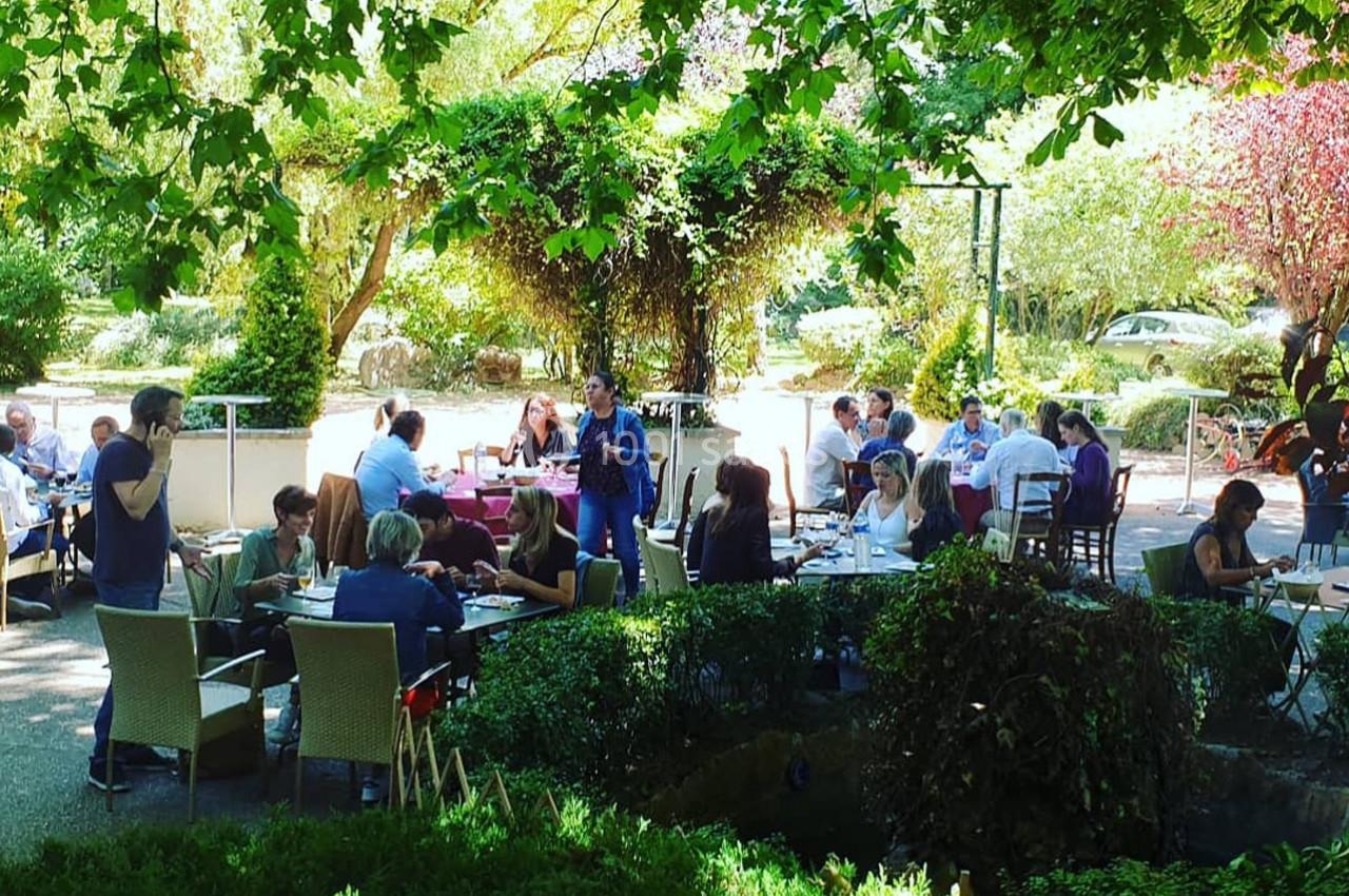 Terrasse ombragée d'un restaurant avec des clients attablés, entourée de verdure et d'arbres en plein été.