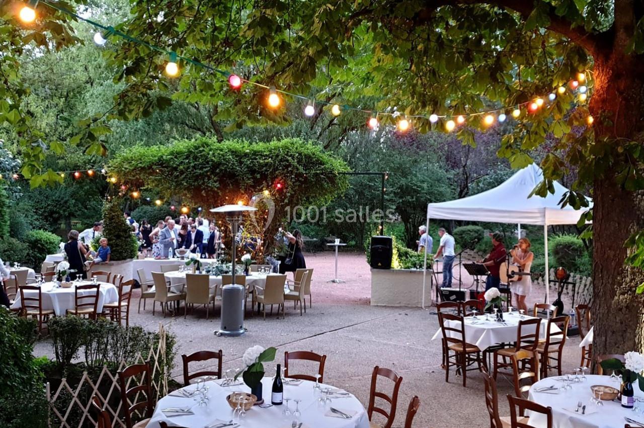 Terrasse extérieure avec tables dressées, guirlandes lumineuses, musiciens et invités dans un cadre verdoyant.
