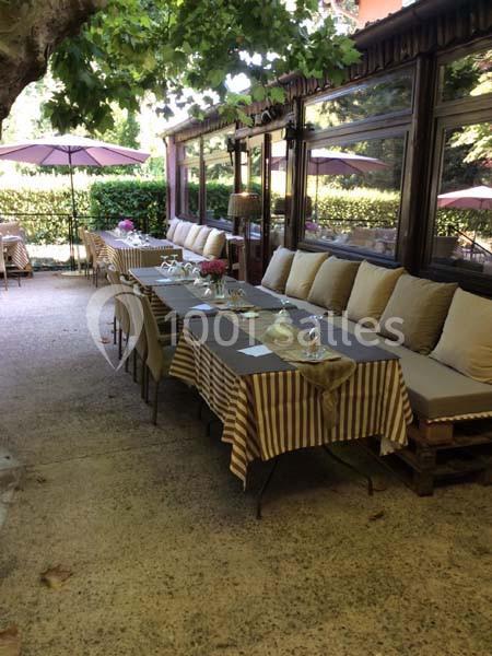 Terrasse ombragée avec tables dressées, nappes rayées et chaises alignées près d'une haie et d'une véranda.