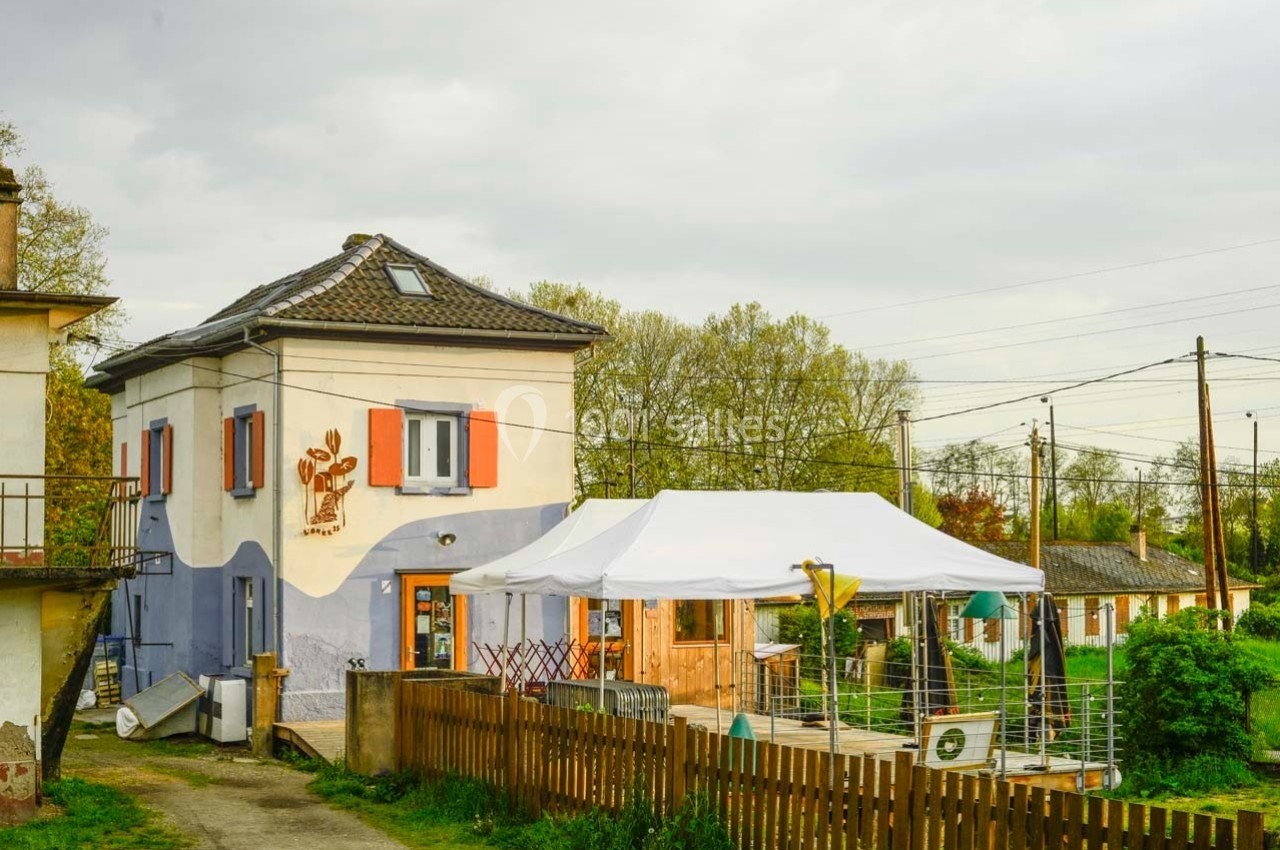 Maison avec volets rouges, terrasse couverte par une tente blanche, entourée de verdure et proche de voies ferrées.