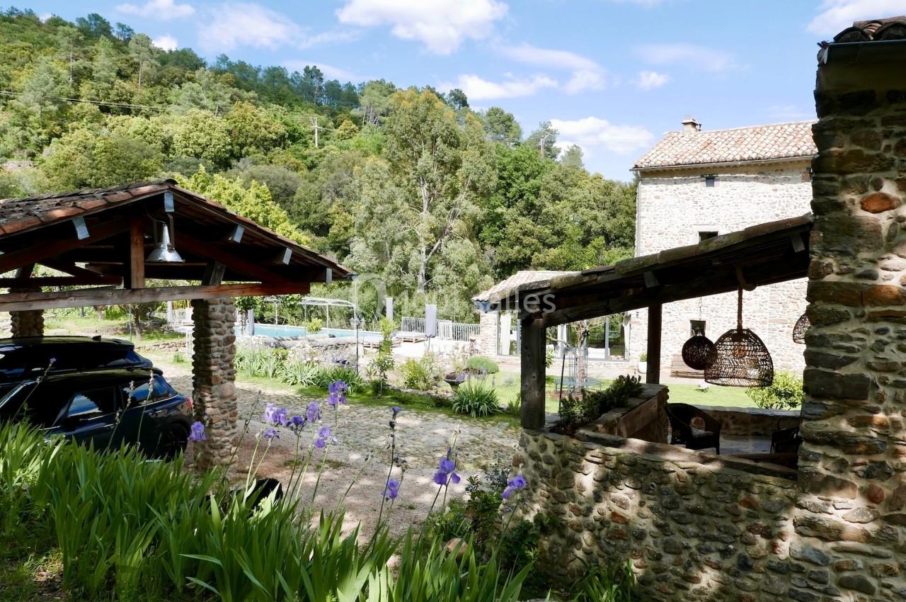 Vue d'une maison en pierre avec terrasse couverte, jardin fleuri et paysage boisé en arrière-plan.