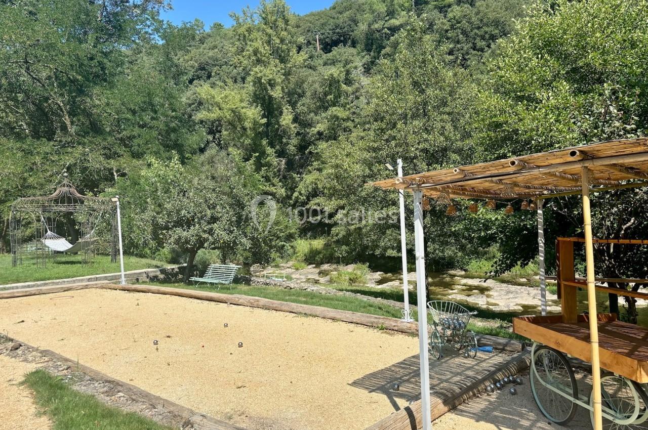 Terrain de pétanque ensoleillé avec pergola en bois, entouré de verdure et d'arbres, près d'une rivière.