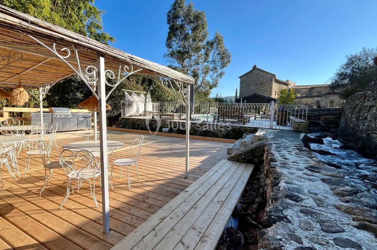 Terrasse en bois avec mobilier en fer forgé, pergola, ruisseau et vue sur une bâtisse en pierre sous un ciel dégagé.