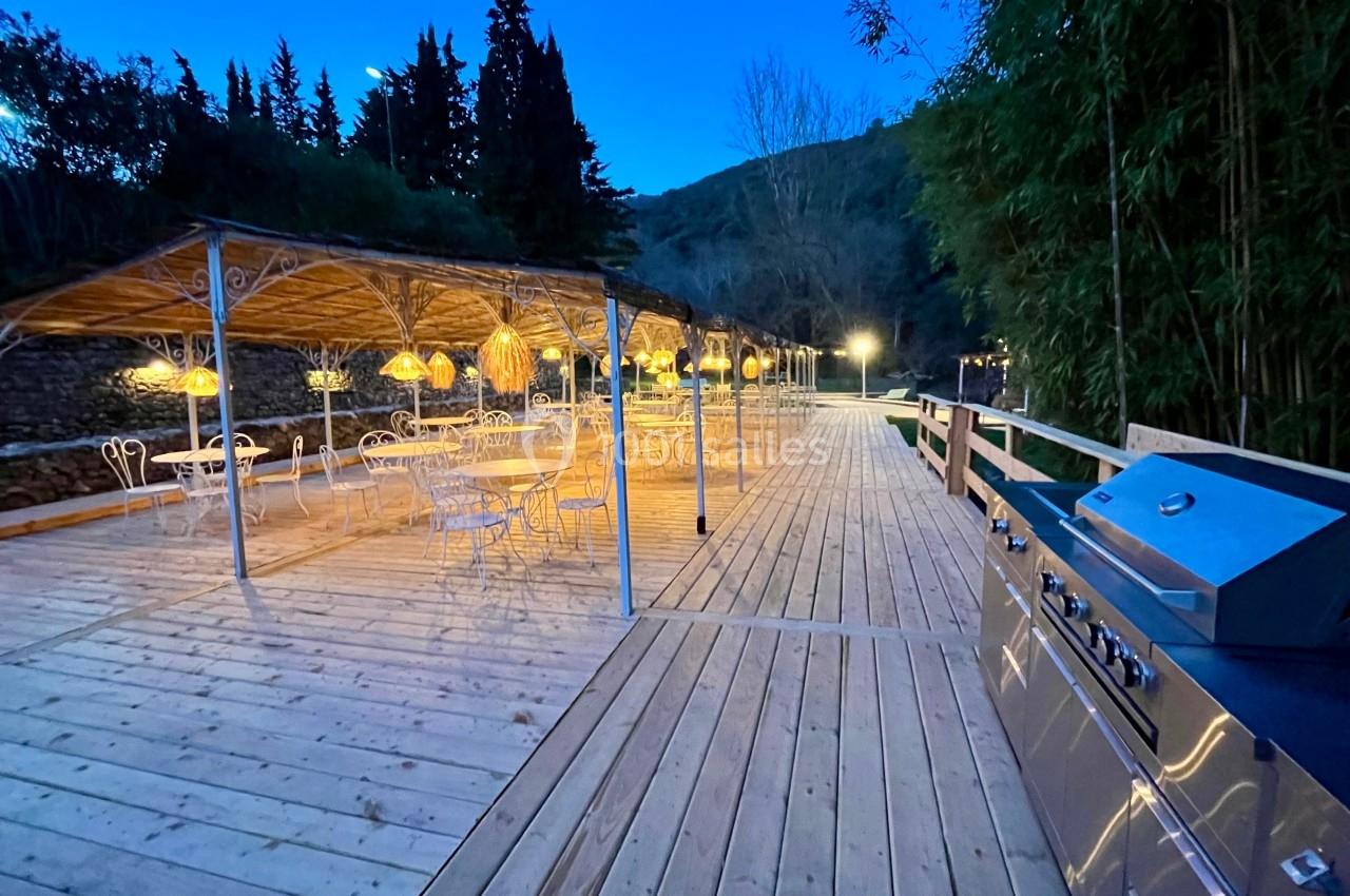 Terrasse en bois éclairée au crépuscule avec tables, chaises, pergolas et un barbecue en premier plan.