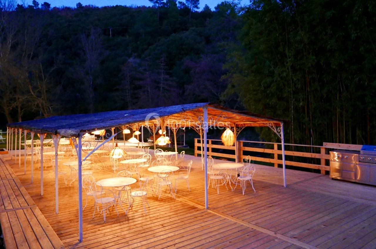 Terrasse en bois éclairée le soir, avec des tables et chaises blanches sous une pergola, entourée de végétation.