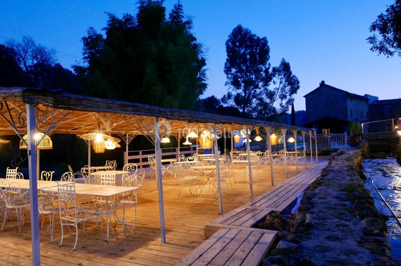 Terrasse en bois éclairée par des lampes suspendues, avec des tables et chaises en métal, entourée de végétation au…