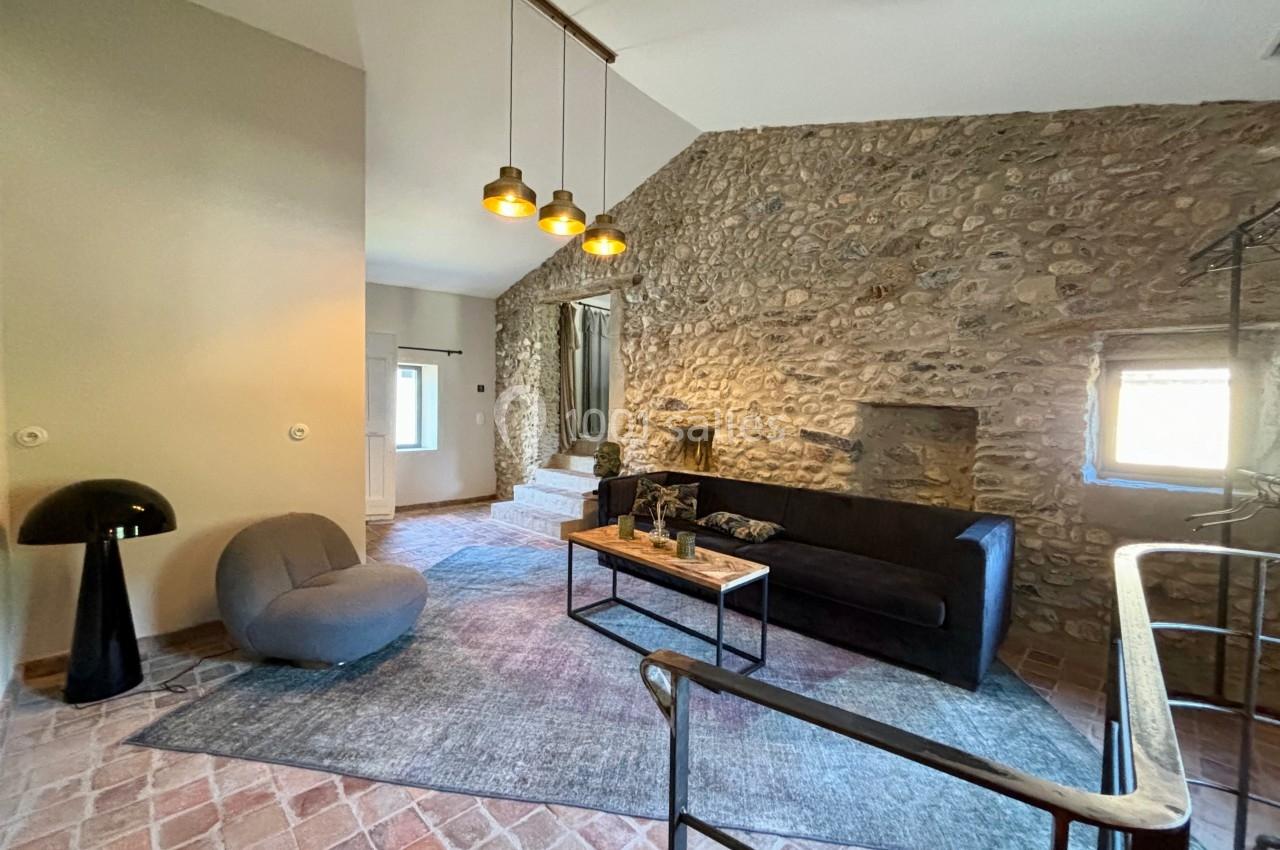 Salon avec murs en pierre, canapé noir, fauteuil gris, tapis bleu, éclairage suspendu et sol en carreaux de terre cuite.