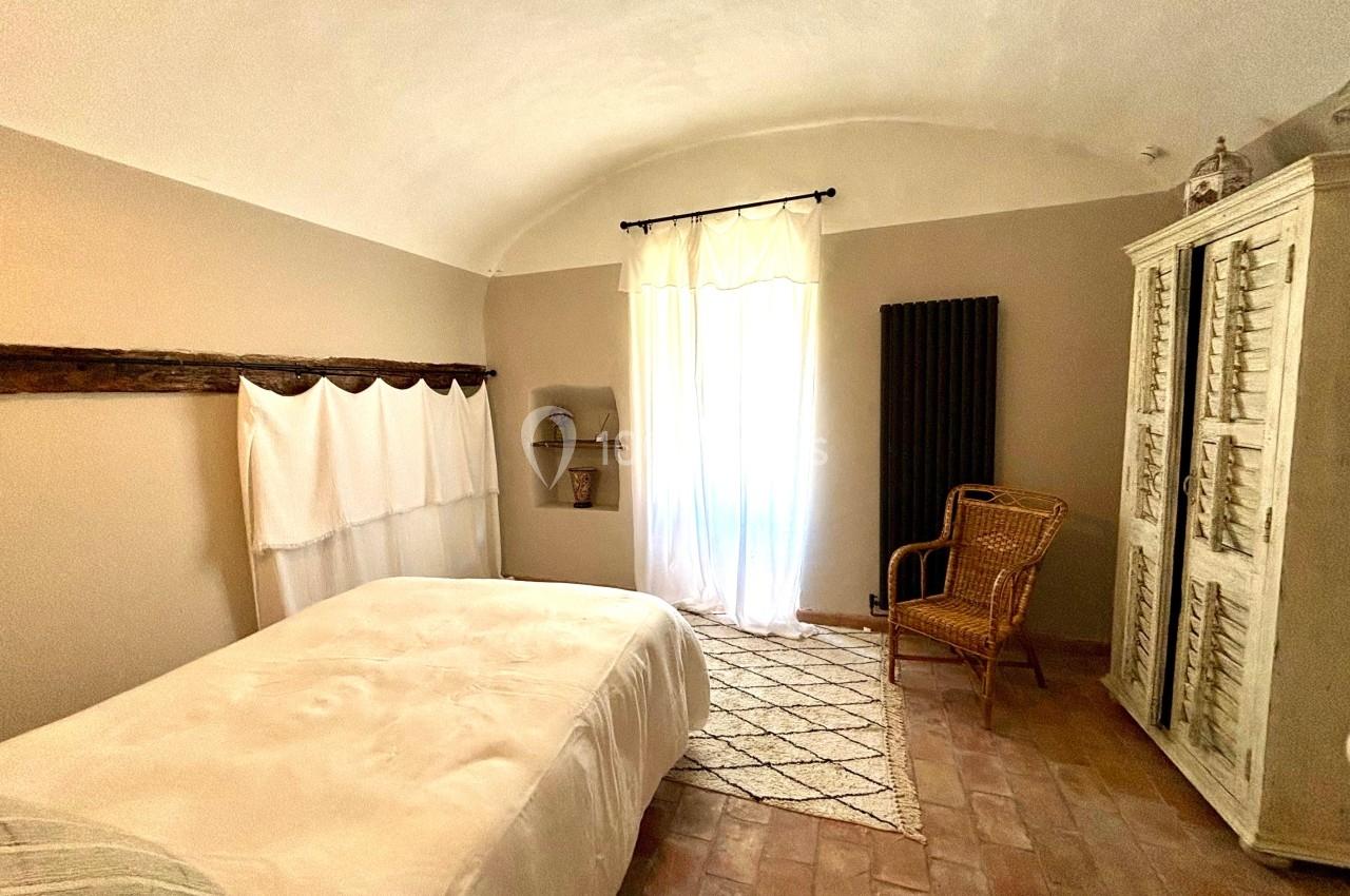 Chambre lumineuse avec lit double, armoire blanche, fauteuil en osier et sol en terre cuite, éclairée par une fenêtre.