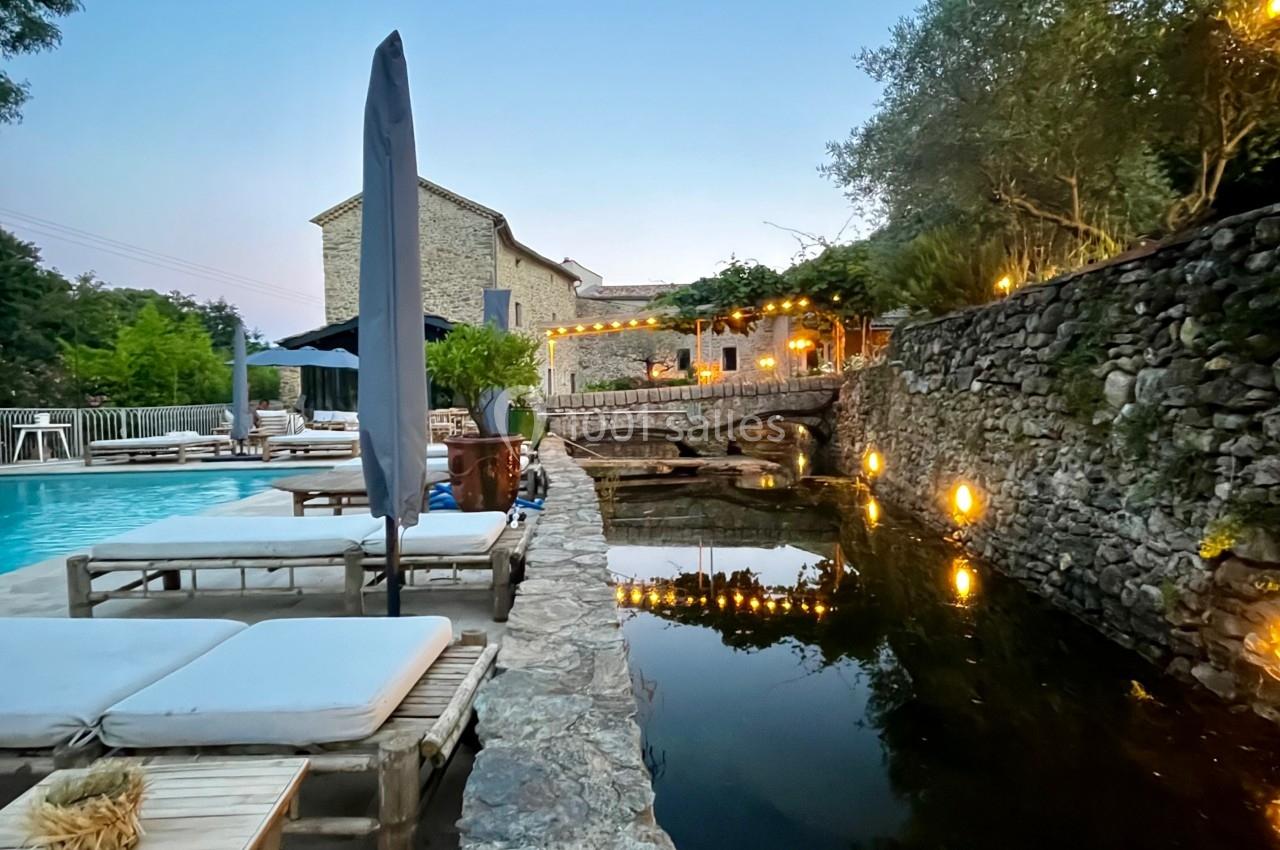 Terrasse avec piscine, chaises longues, parasol et bassin d'eau entouré de pierres, éclairée à la tombée de la nuit.