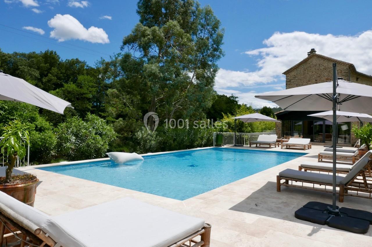 Piscine extérieure entourée de transats et parasols, avec végétation et maison en pierre en arrière-plan.
