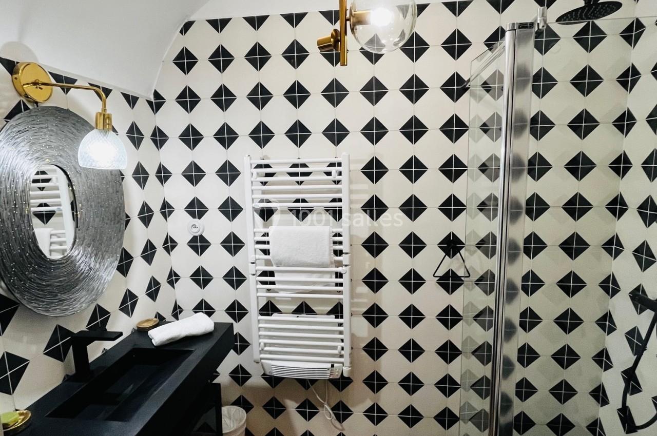Salle de bain moderne avec carrelage géométrique noir et blanc, douche vitrée, lavabo noir et miroir rond texturé.