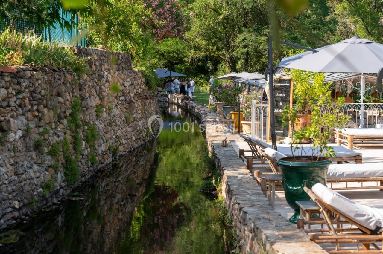Canal bordé de pierres avec des chaises longues et parasols, entouré de végétation et d'un jardin arboré.