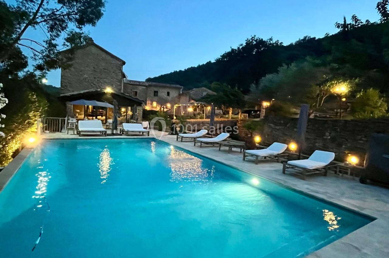 Piscine éclairée au crépuscule entourée de chaises longues, avec des maisons en pierre et végétation en arrière-plan.