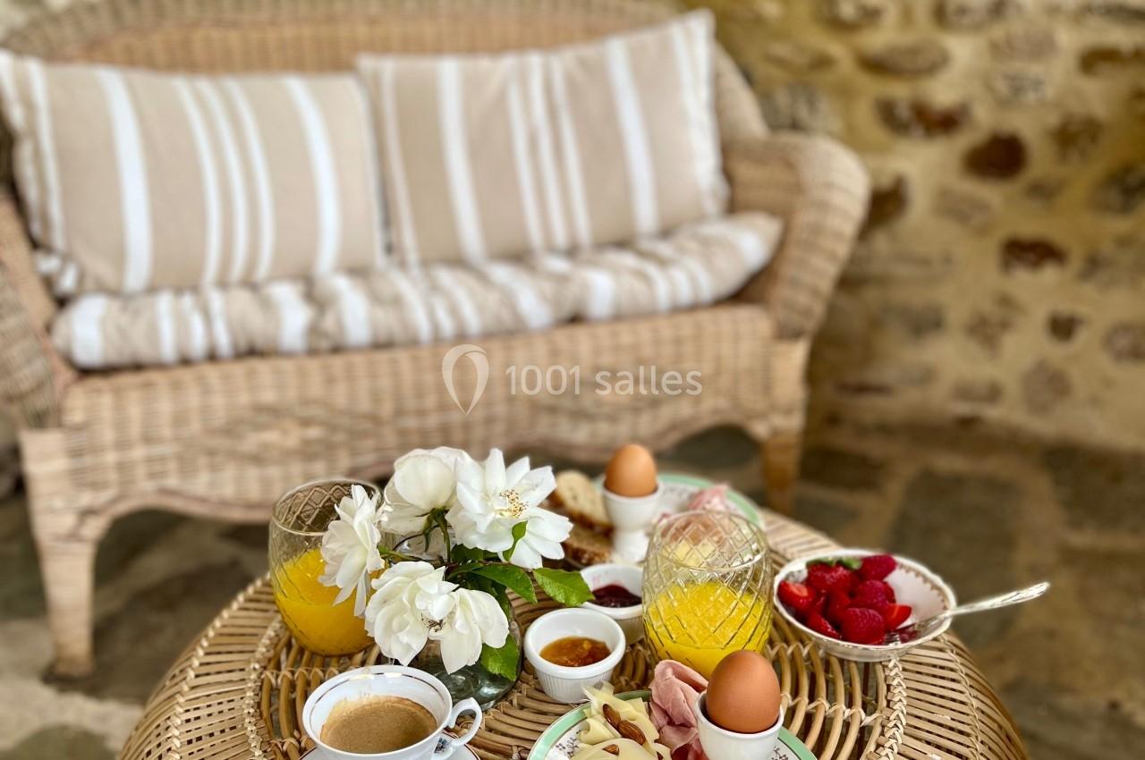 Petit-déjeuner composé de pain, charcuterie, œufs, fruits et café, servi sur une table en rotin dans un cadre champêtre.