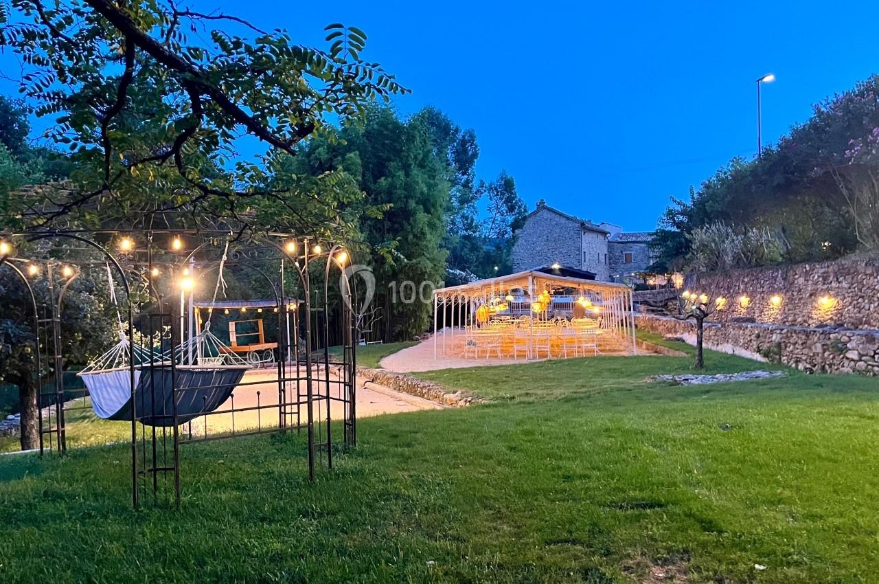 Jardin éclairé au crépuscule avec un hamac suspendu, une pergola lumineuse et des tables dressées en arrière-plan.