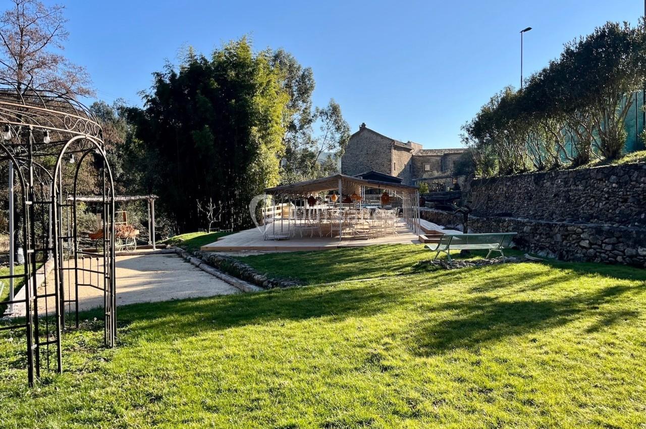 Jardin avec pelouse, pergola métallique, terrasse en bois et bâtiment en pierre sous un ciel dégagé.