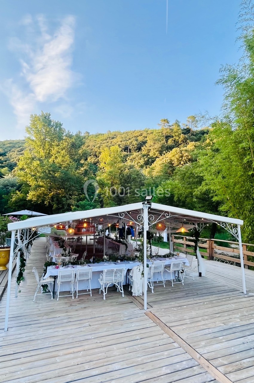 Pergola blanche décorée, installée sur une terrasse en bois, entourée de verdure et éclairée par des guirlandes lumineuses.