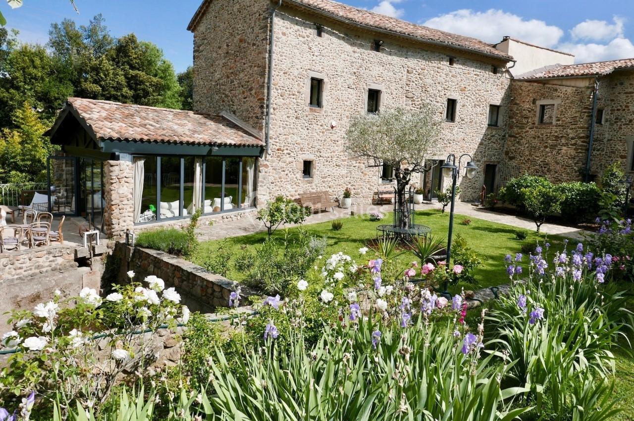 Jardin fleuri avec pelouse, arbres et terrasse, entouré d'un bâtiment en pierre sous un ciel dégagé.
