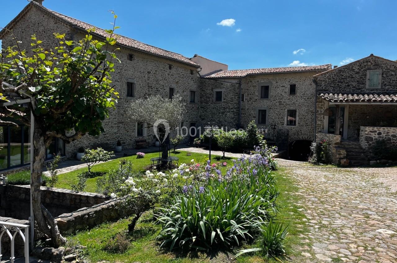 Cour intérieure d'une maison en pierre avec jardin fleuri, arbres, allée pavée et ciel bleu en arrière-plan.