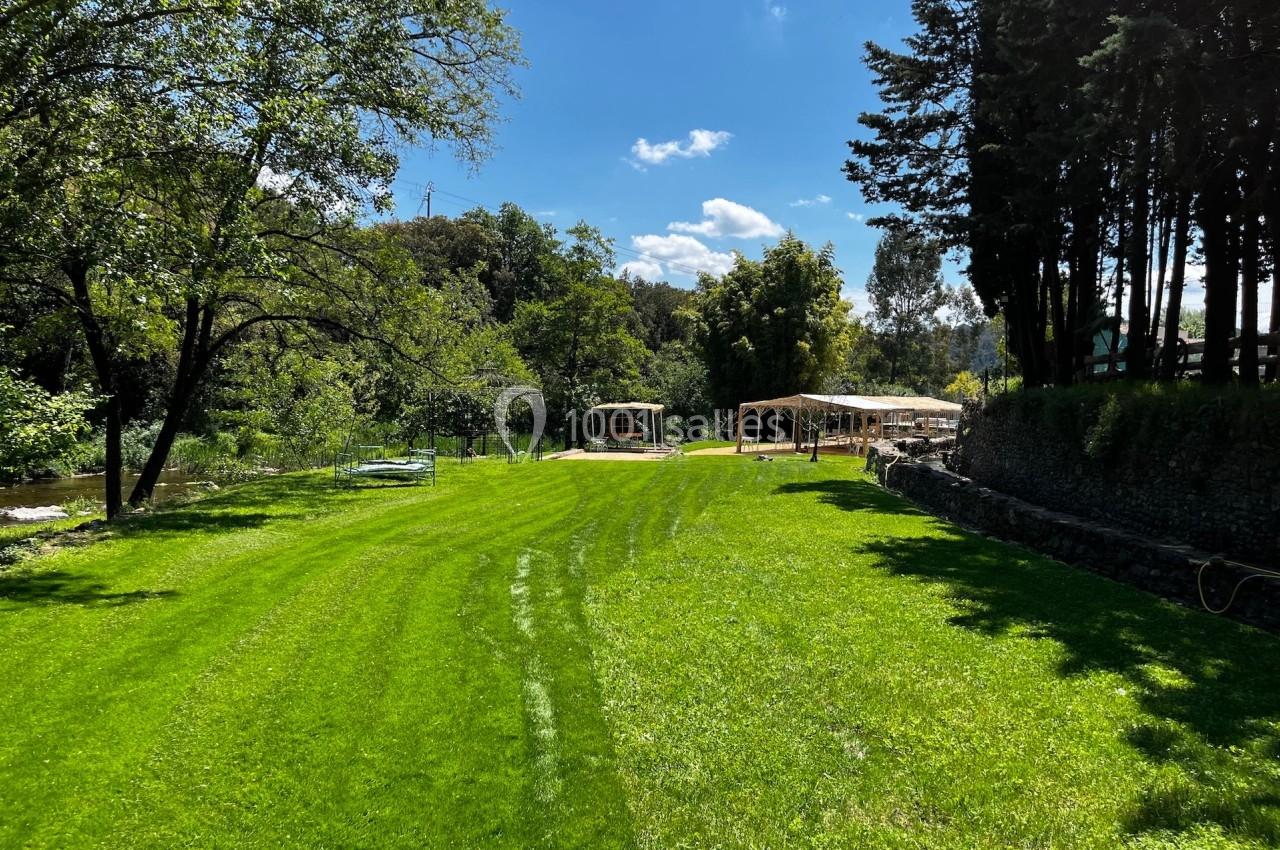 Pelouse verdoyante entourée d'arbres, avec des structures en bois et un ciel bleu en arrière-plan.