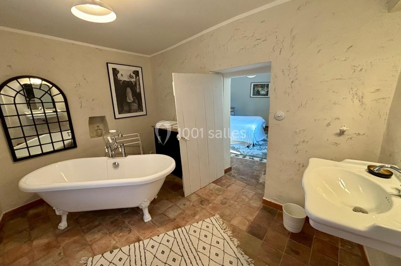 Salle de bain avec baignoire sur pieds, lavabo, miroir, tapis beige et porte ouverte sur une chambre adjacente.