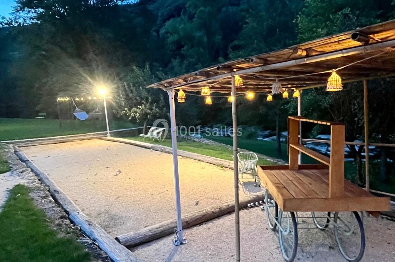 Terrain de pétanque éclairé en soirée, avec une pergola en bois et des guirlandes lumineuses dans un jardin verdoyant.