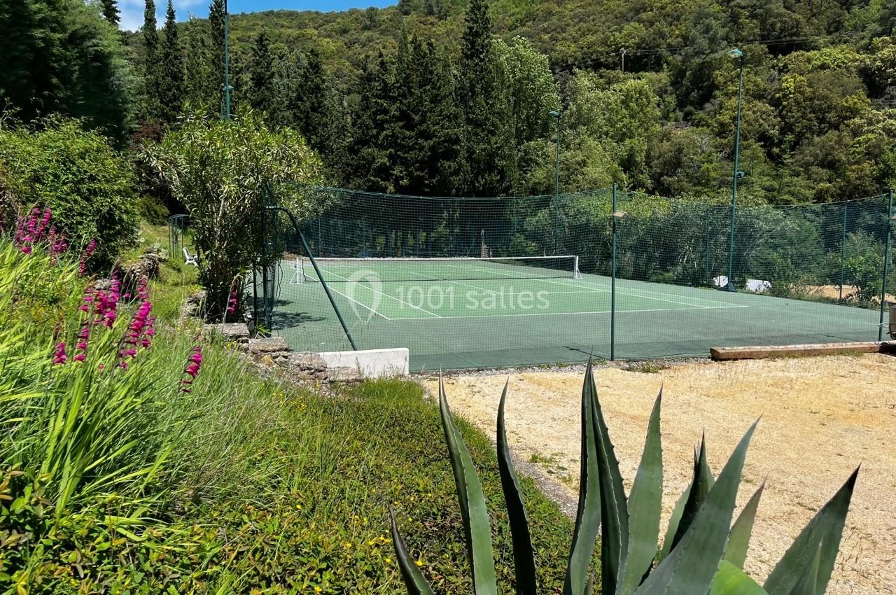 Terrain de tennis extérieur entouré de végétation, avec un ciel bleu et des arbres en arrière-plan.