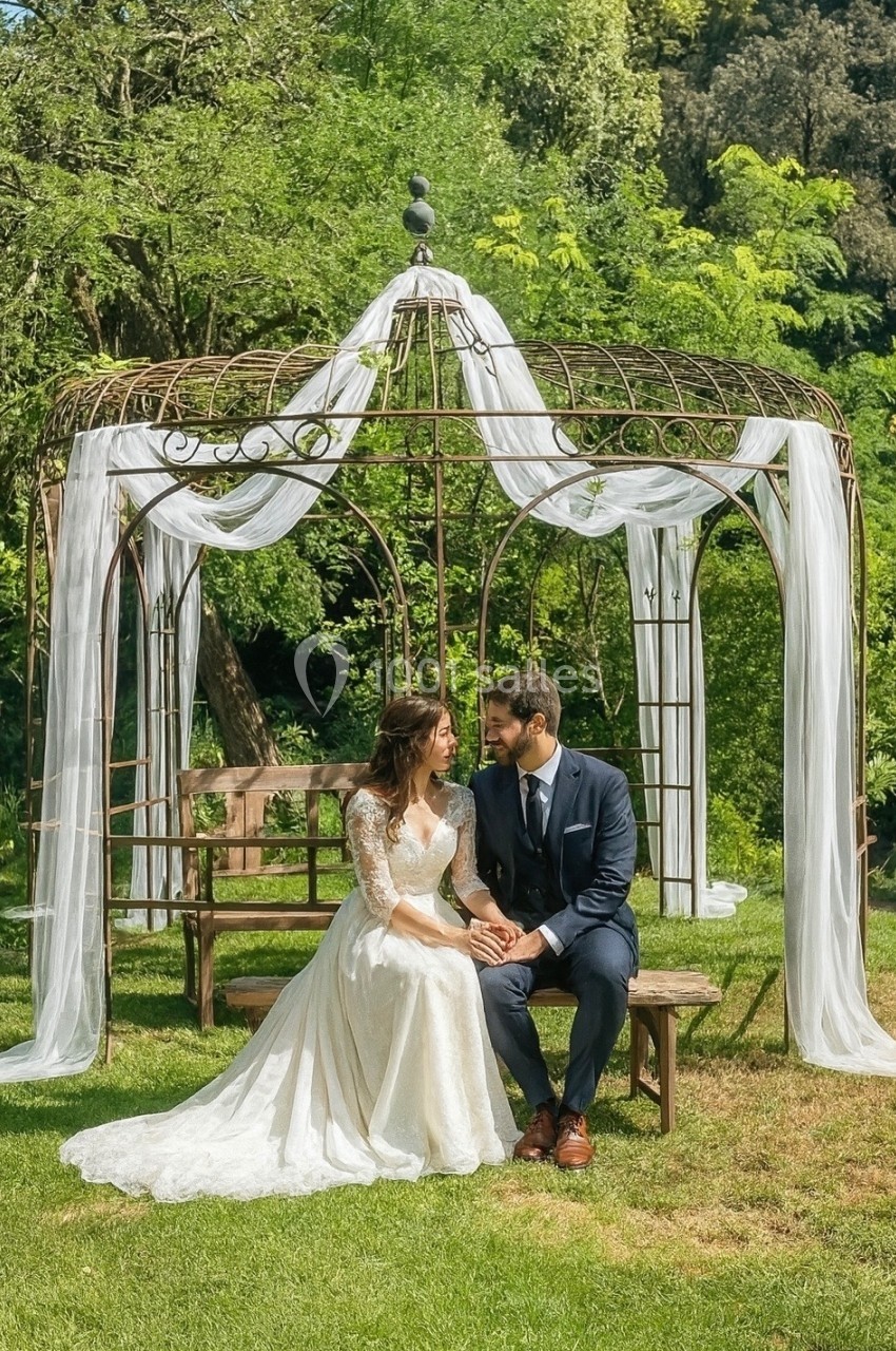 Un couple assis sous une pergola décorée de voiles blancs, entourée de verdure dans un jardin.