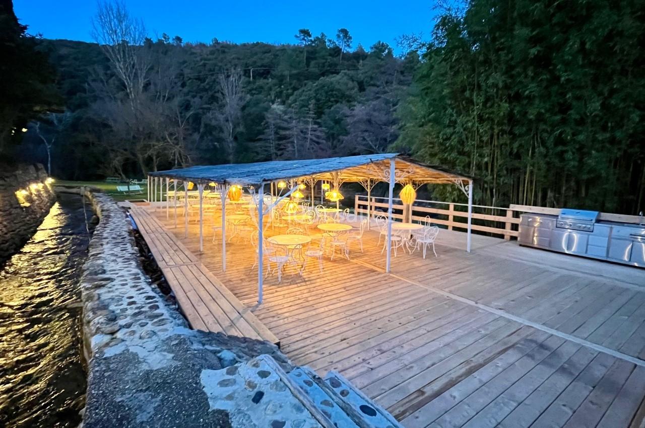 Terrasse en bois éclairée avec des tables et des chaises, située près d'un ruisseau et entourée de végétation.