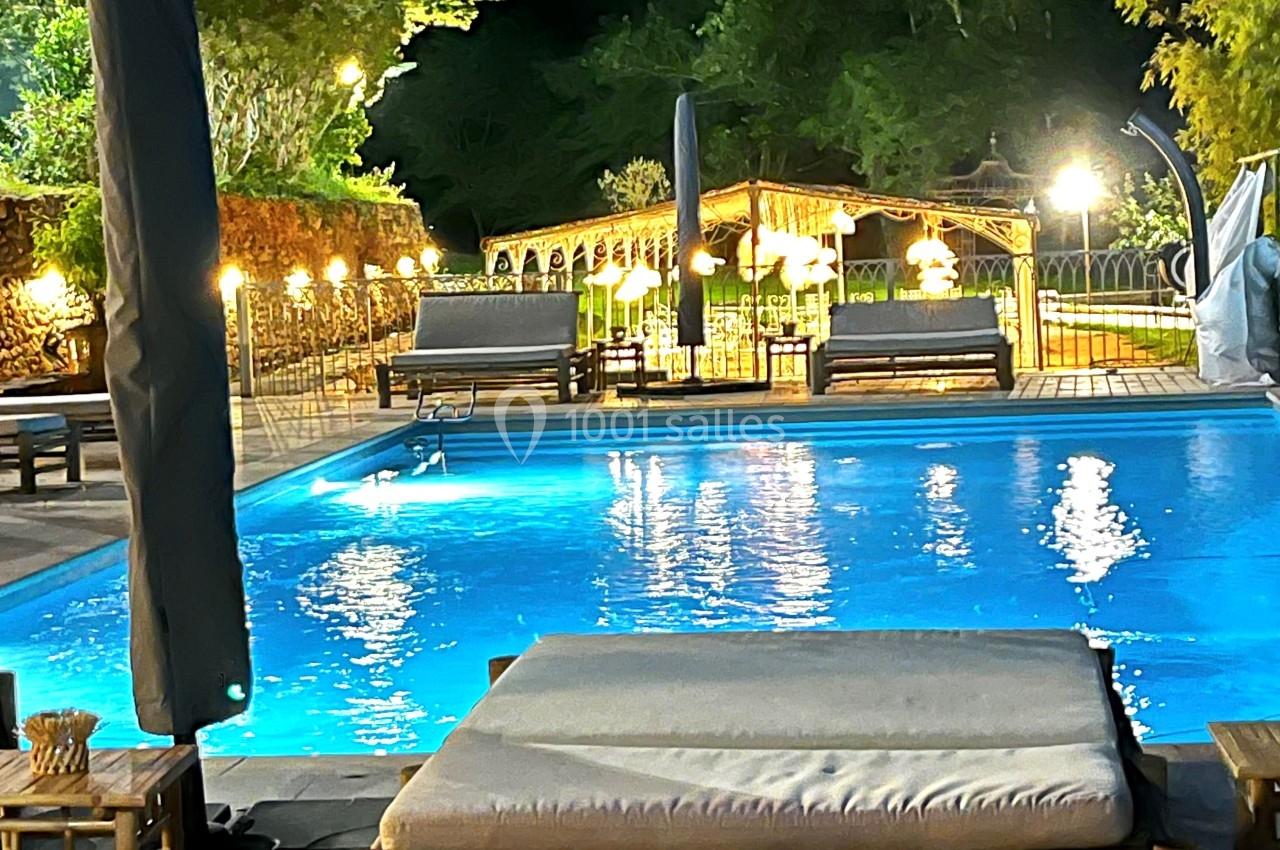 Piscine éclairée de nuit entourée de chaises longues, parasols fermés et végétation en arrière-plan.